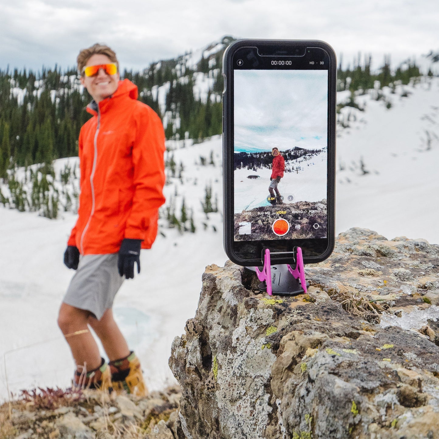 Mann in roter Jacke posiert für iPhone auf Stativ, schneebedeckte Berge im Hintergrund.