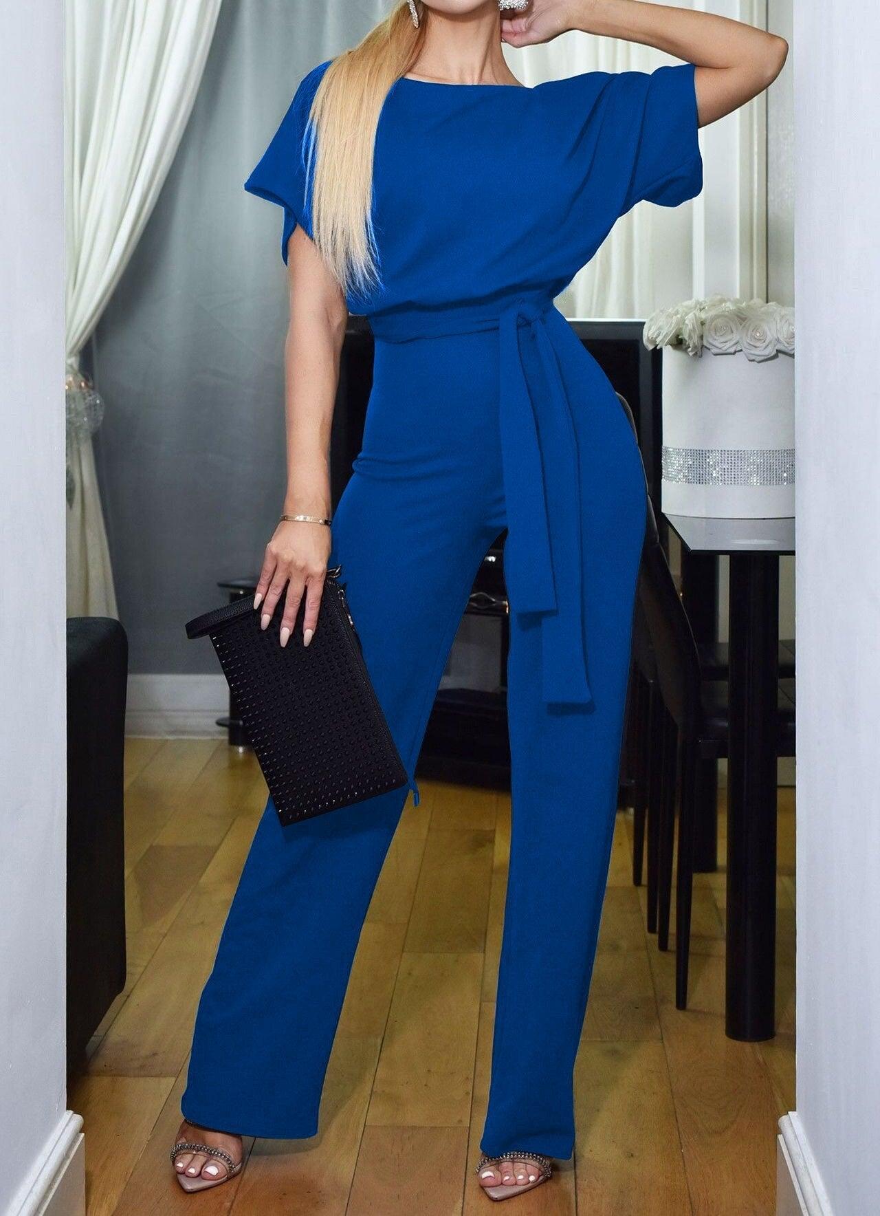 Frau in blauem Jumpsuit mit Gürtel, hält schwarze Clutch, elegantes Abendoutfit, Holzboden.