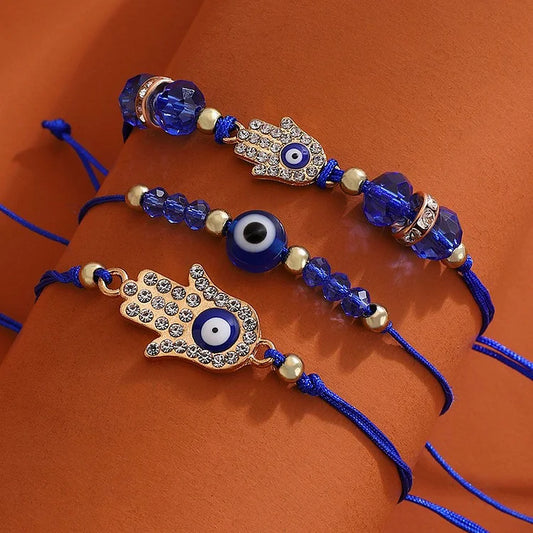 Drei blaue Armbänder mit Hamsa-Hand und Nazar-Amulett, verziert mit Kristallen auf orangefarbenem Hintergrund.