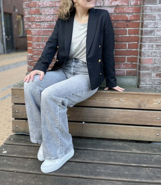 Frau in schwarzem Blazer und grauer Jeans sitzt auf Bank vor Backsteinwand.