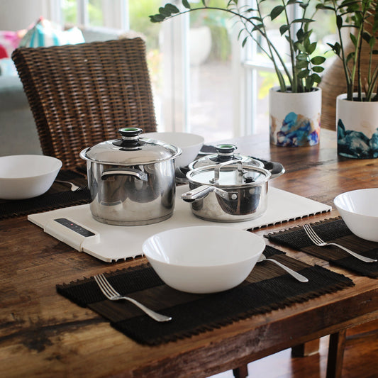 Edelstahl Kochtöpfe auf Holzbrett, Esstisch, weiße Schalen, Pflanzen im Hintergrund.