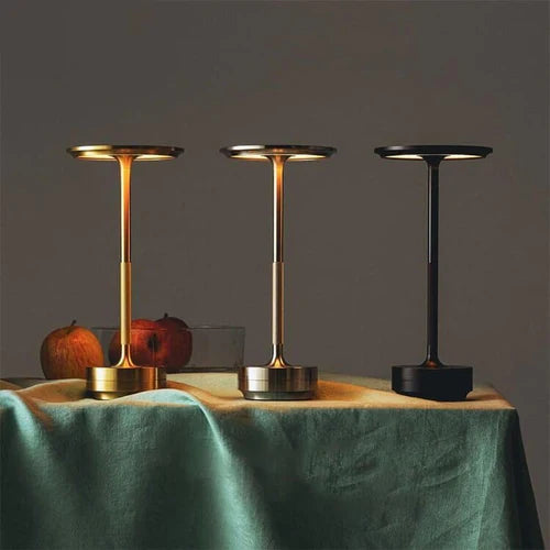 Drei moderne Tischleuchten in Gold und Schwarz auf Tisch mit grüner Decke und Äpfeln.