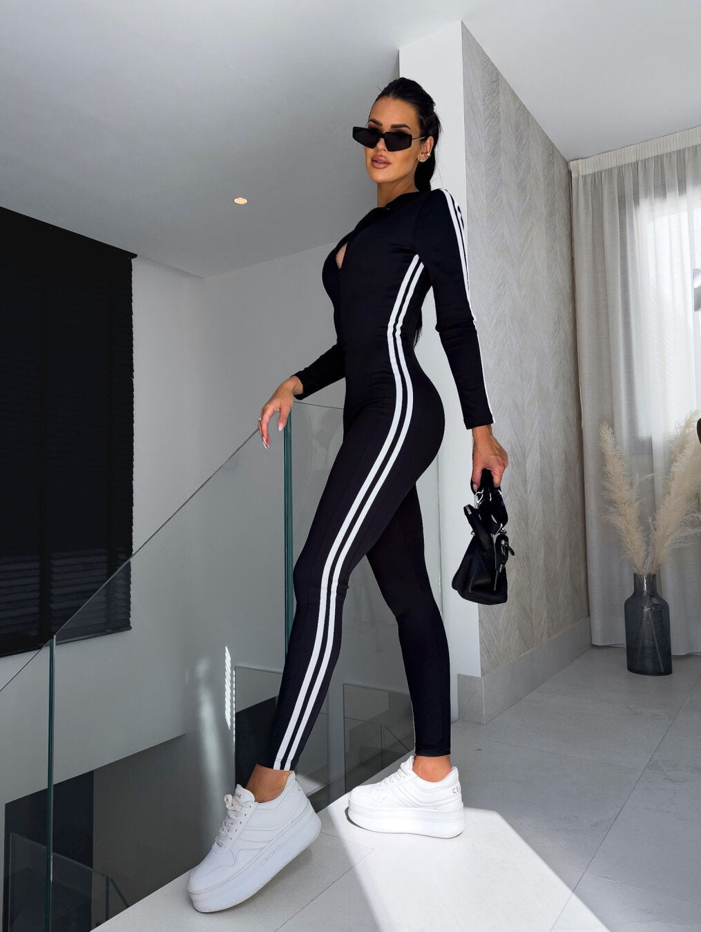 Frau in schwarzem Sportanzug mit weißen Streifen, trägt Sonnenbrille und weiße Sneaker.