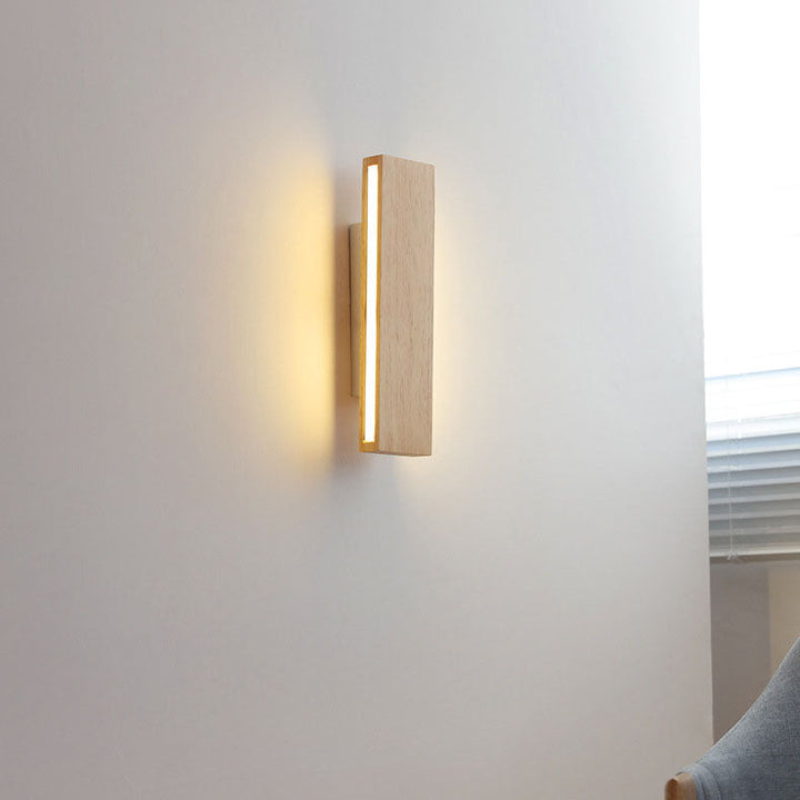 Rozsvícená svislá dřevěná lampa na bílé zdi u okna se žaluziemi, vedle šedé opěradlo křesla. 15