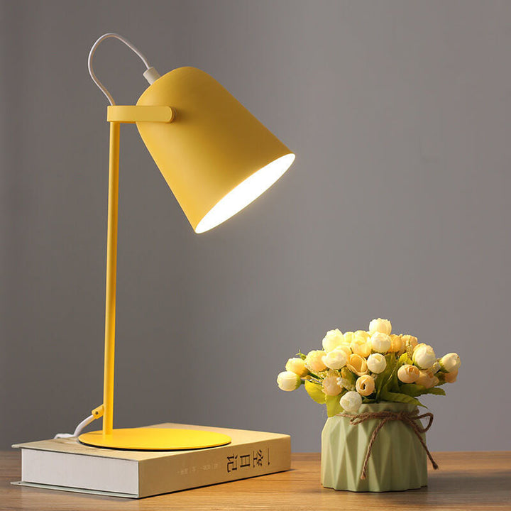 Žlutá kovová stolní lampa se svítícím kuželovým stínidlem stojí na knize vedle kytice bílých a krémových poupat ve zeleném květináči. 1