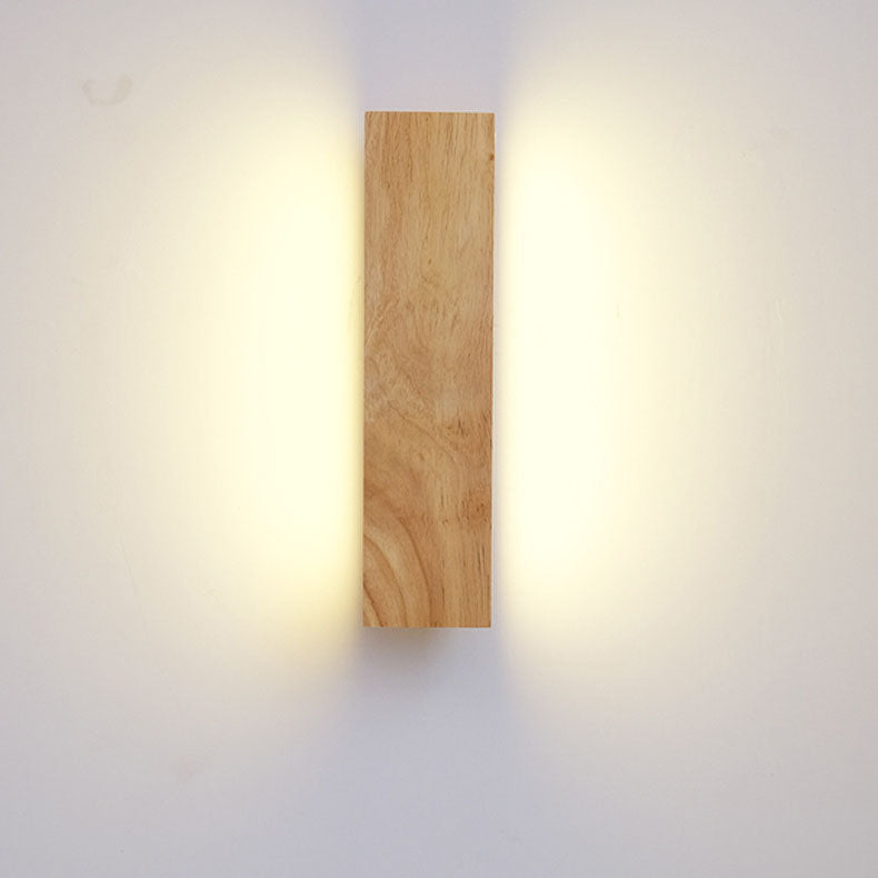 Svisle zavěšená dřevěná lampa zepředu, z obou boků vyzařuje teplé světlo na světlou zeď. 3