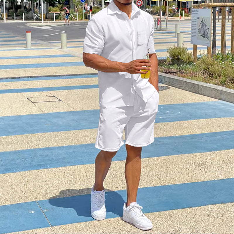 Mann in weißem Sommer-Shorts-Set, hält Getränk, trägt weiße Sneaker, urbaner Hintergrund.