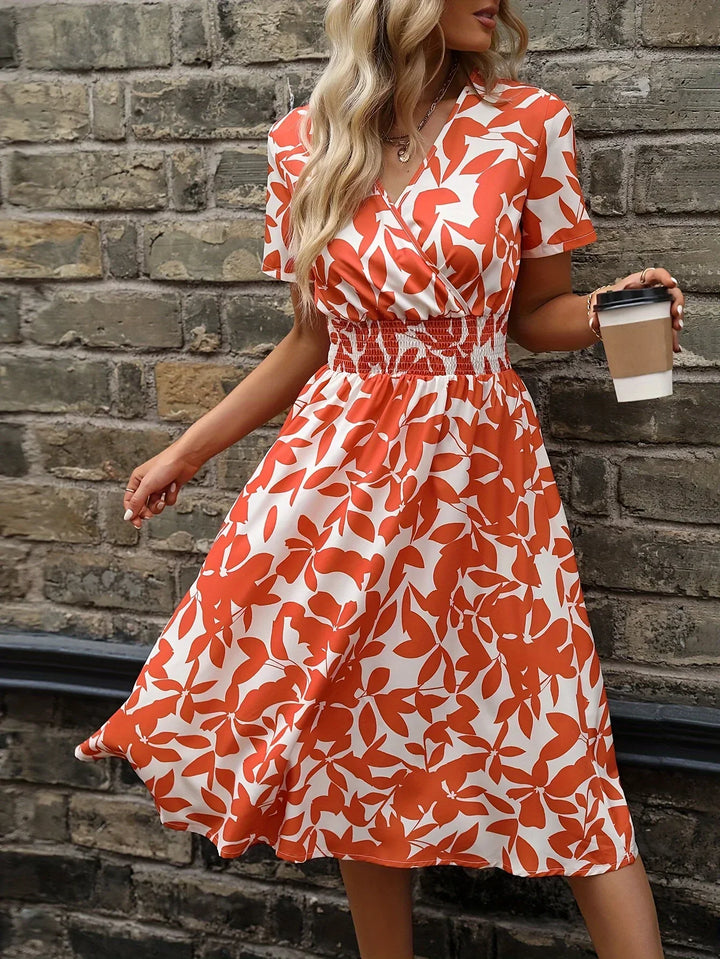 Frau in rotem Sommerkleid mit Blattmuster hält Kaffeebecher vor Backsteinmauer.