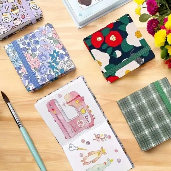 Notizbücher mit bunten Stoffeinbänden auf Holztisch, Blumenmuster, Aquarellpinsel, kreative Schreibwaren.