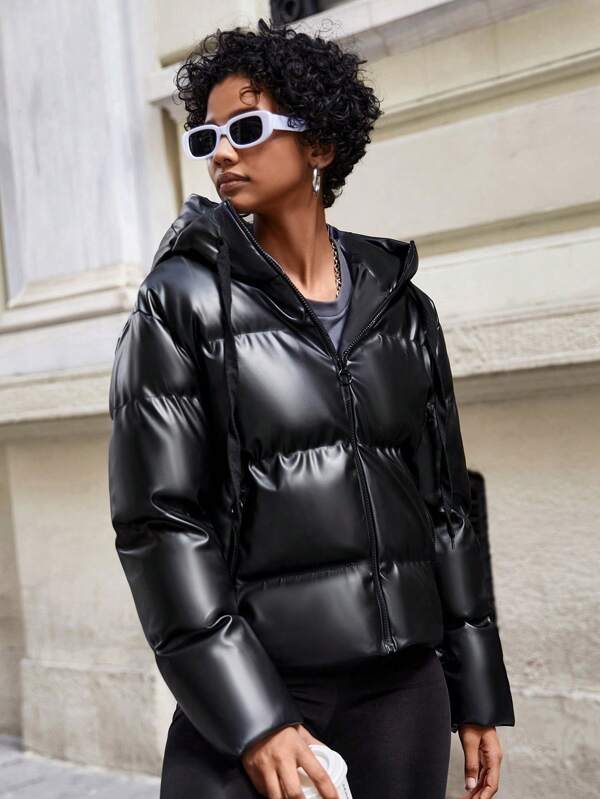 Frau in schwarzer, glänzender Daunenjacke mit Kapuze und Sonnenbrille auf städtischer Straße.