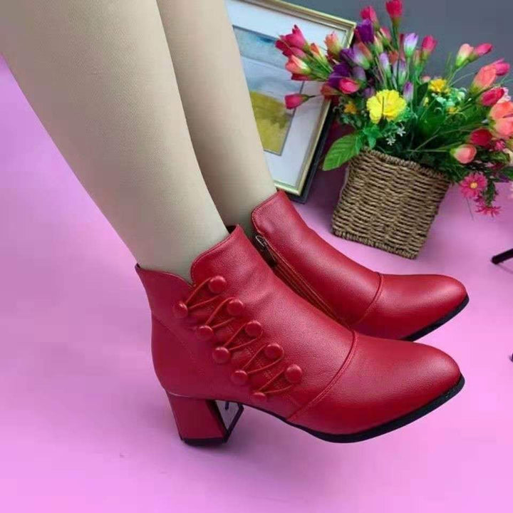 Rote Damenstiefeletten mit Absatz, Lederoptik, dekorative Knöpfe, Blumen im Hintergrund.