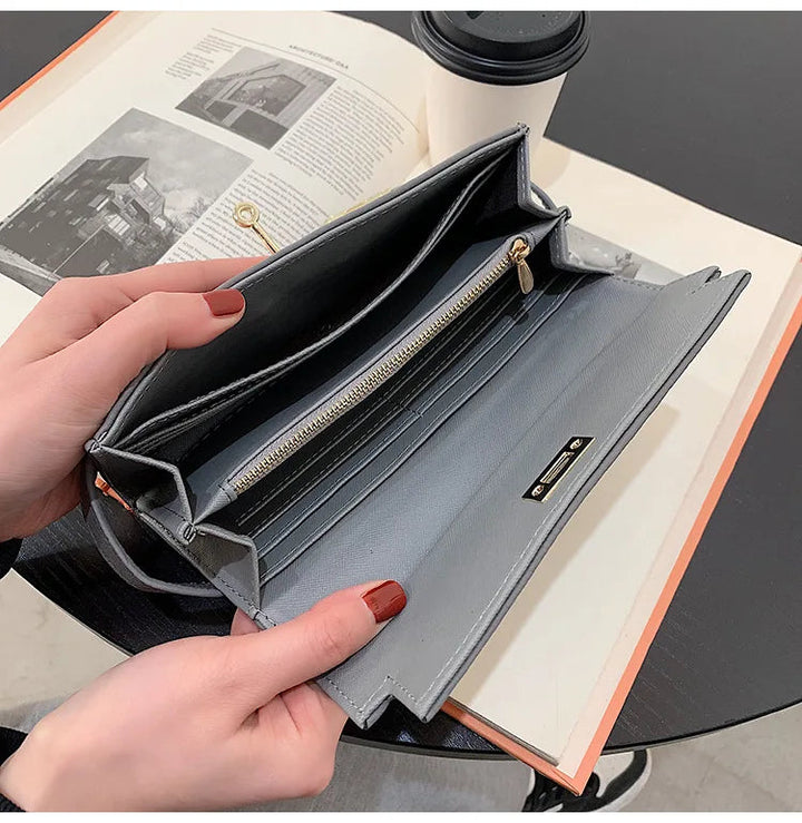 Graue Damen-Geldbörse aus Leder mit Reißverschluss, geöffnet auf Tisch mit Buch und Kaffeetasse.