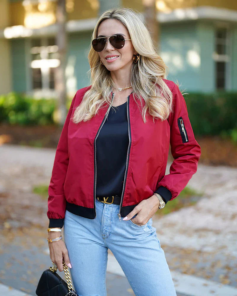 Frau in roter Bomberjacke, schwarze Sonnenbrille, blaue Jeans, draußen, lässiger Stil.
