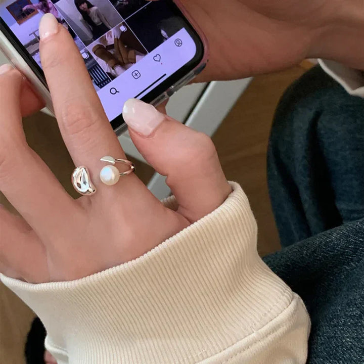 Hand mit Smartphone, silbernem Ring und Perlenring, Instagram-Feed, beige Pullover, Jeans.
