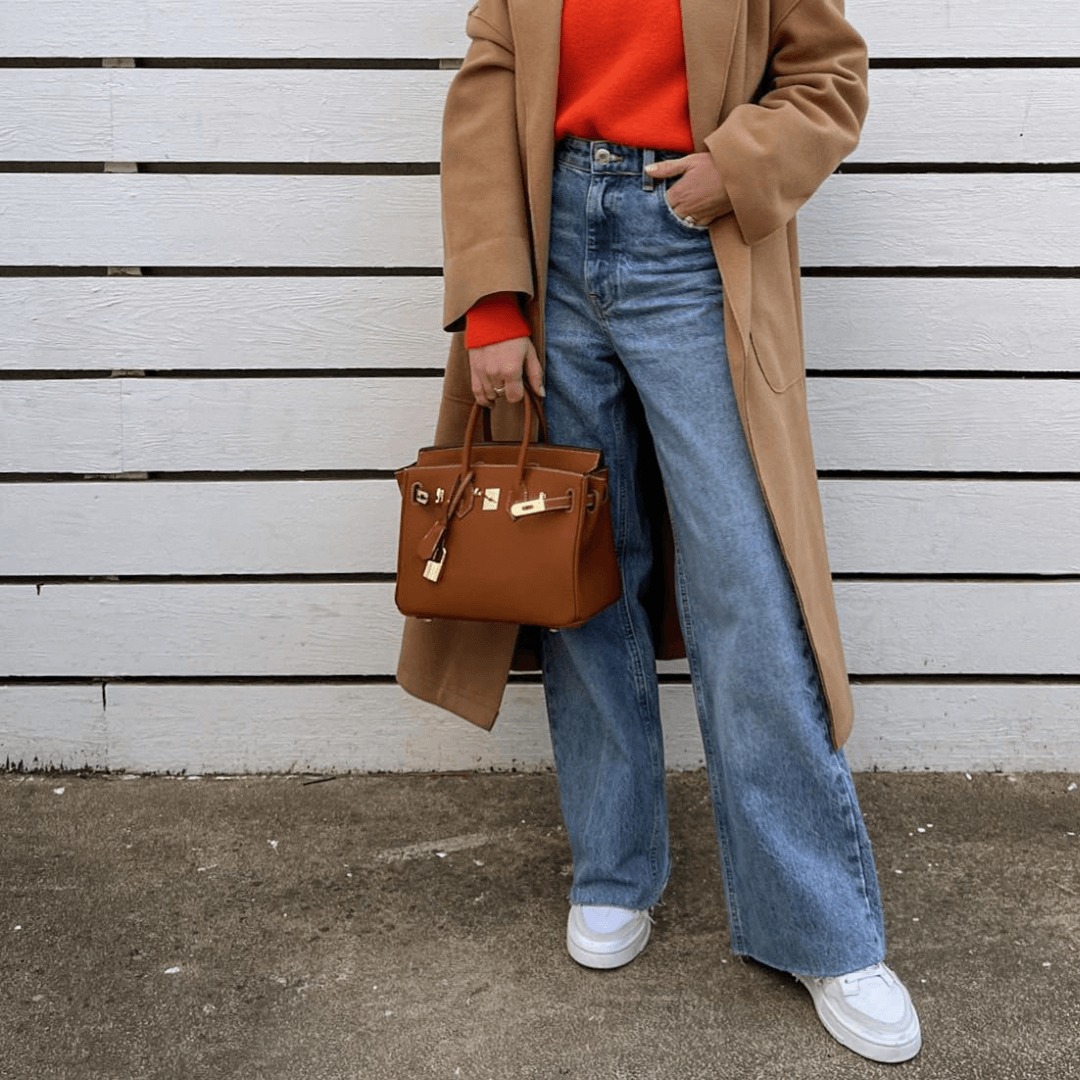 Frau in beigem Mantel, rotem Pullover, blauer Jeans, weißem Sneaker, brauner Handtasche.