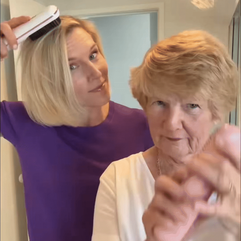 Blonďatá žena v fialovém triku si přikládá bílý kartáč k vlasům, před ní stojí starší dáma s krátkým účesem. 9