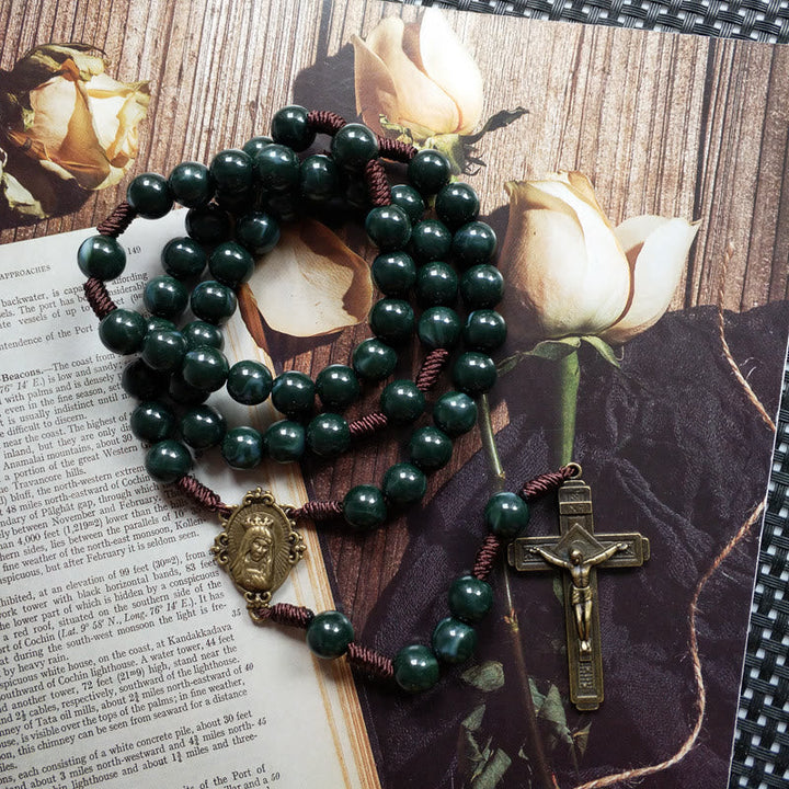 Dunkelgrüner Rosenkranz mit Kreuz und Buddha-Anhänger auf Buch und Rosenhintergrund.
