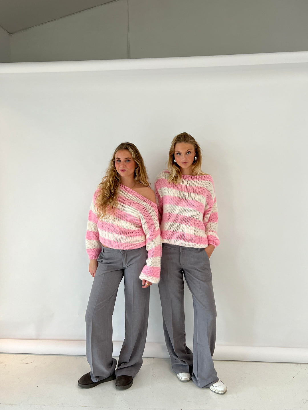 Zwei Frauen in gestreiften rosa Pullovern und grauen Hosen vor weißem Hintergrund.