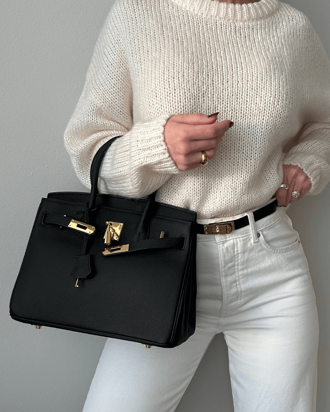 Frau mit schwarzer Lederhandtasche, goldene Details, cremefarbener Pullover, weiße Hose.