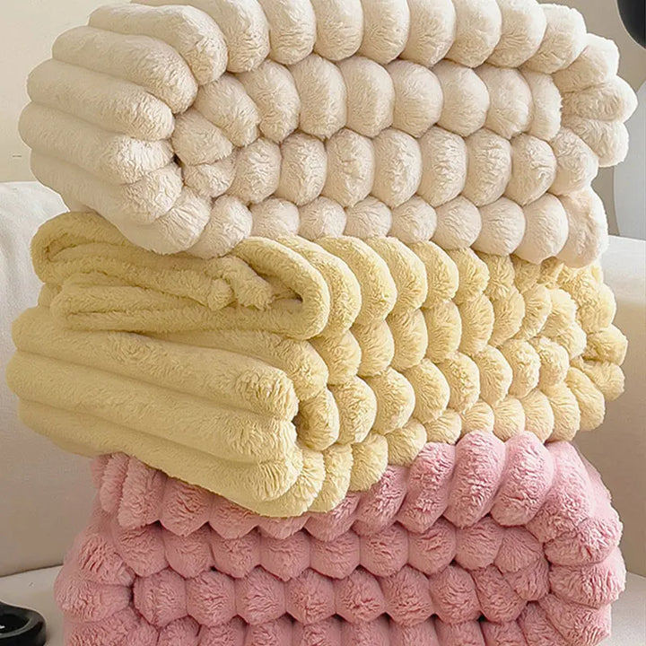 Drei gefaltete, flauschige Decken in Creme, Gelb und Rosa auf einem Sofa gestapelt.