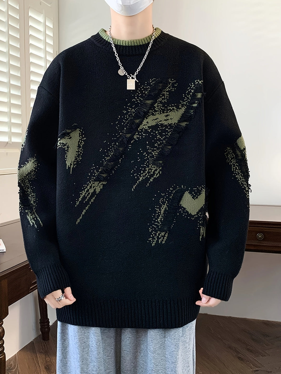 Person trägt schwarzen Strickpullover mit grünem Muster, Kette und grauer Hose.
