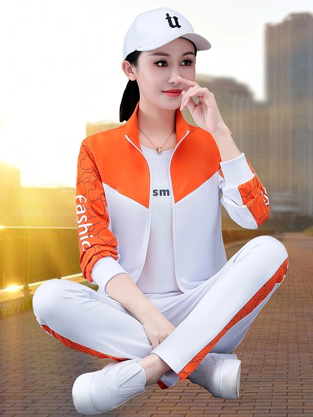 Frau in weißem und orangefarbenem Sportanzug, Baseballkappe, sitzt im Freien, Stadt im Hintergrund.