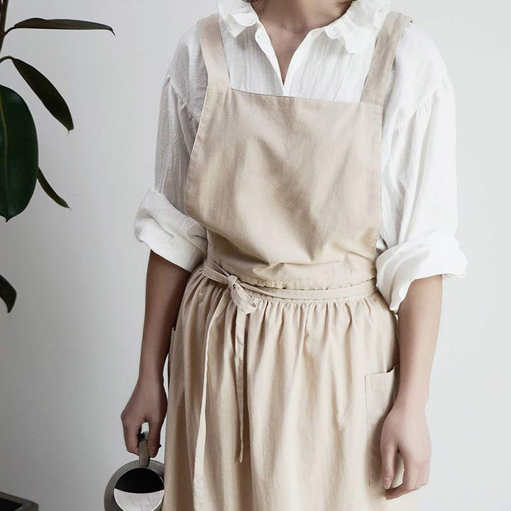Frau in beigem Leinenkleid mit weißer Bluse, hält eine Gießkanne, minimalistisch, modern.