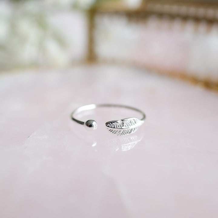 Silberner offener Ring mit Federdesign auf rosa Hintergrund, minimalistischer Schmuck.