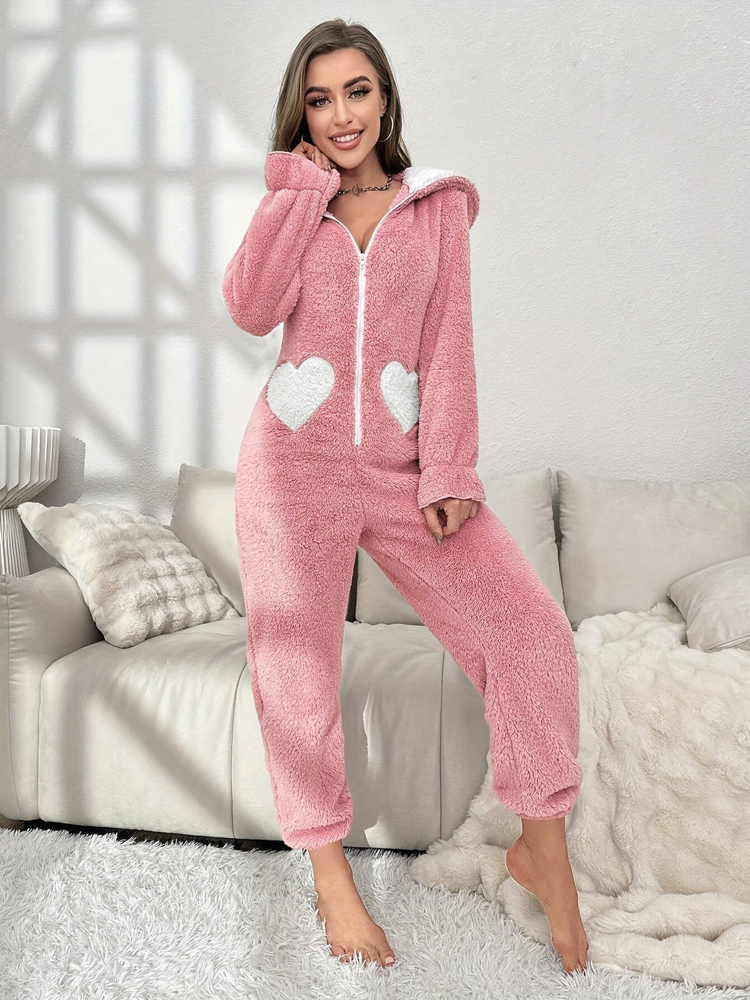 Frau in rosa Fleece-Overall mit Herzmotiven, gemütlich auf Sofa, Wintermode, Damenbekleidung.