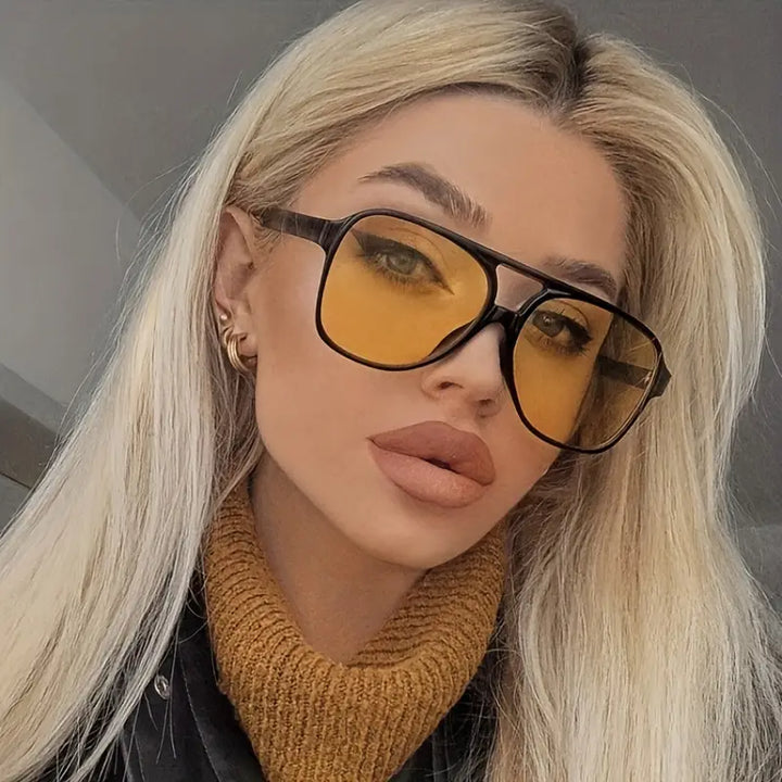 Blonde Frau mit gelber Sonnenbrille, schwarzer Jacke und braunem Rollkragenpullover.