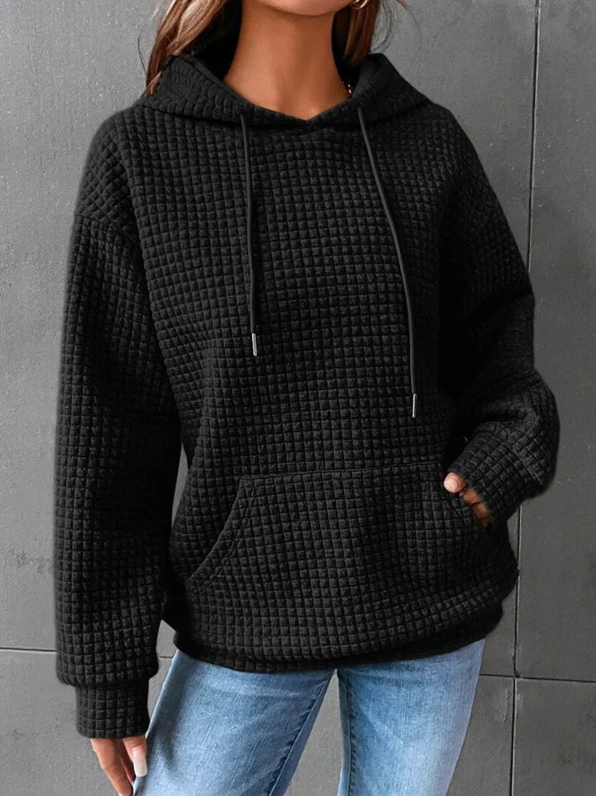 Frau in schwarzem Kapuzenpullover mit Waffelmuster und blauer Jeans vor grauer Wand.