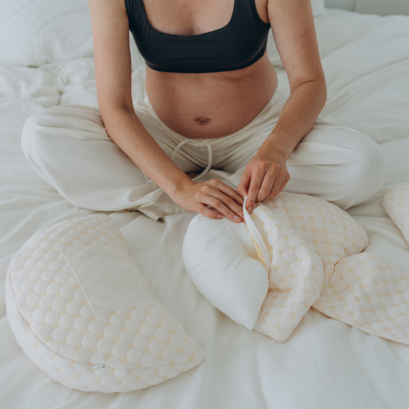 Schwangere Frau mit Stillkissen-Set auf weißem Bett, beige Bezug, bequemes Stillzubehör.