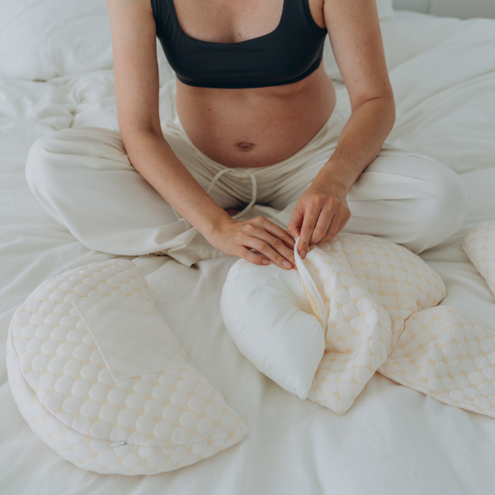 Schwangere Frau mit Stillkissen-Set auf weißem Bett, beige Bezug, bequemes Stillzubehör.