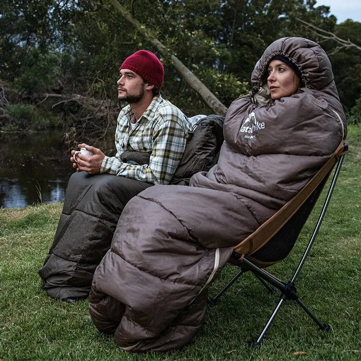 Zwei Personen in Schlafsäcken, Campingstuhl, Outdoor, Natur, Flussufer, Abend, Freizeitkleidung.
