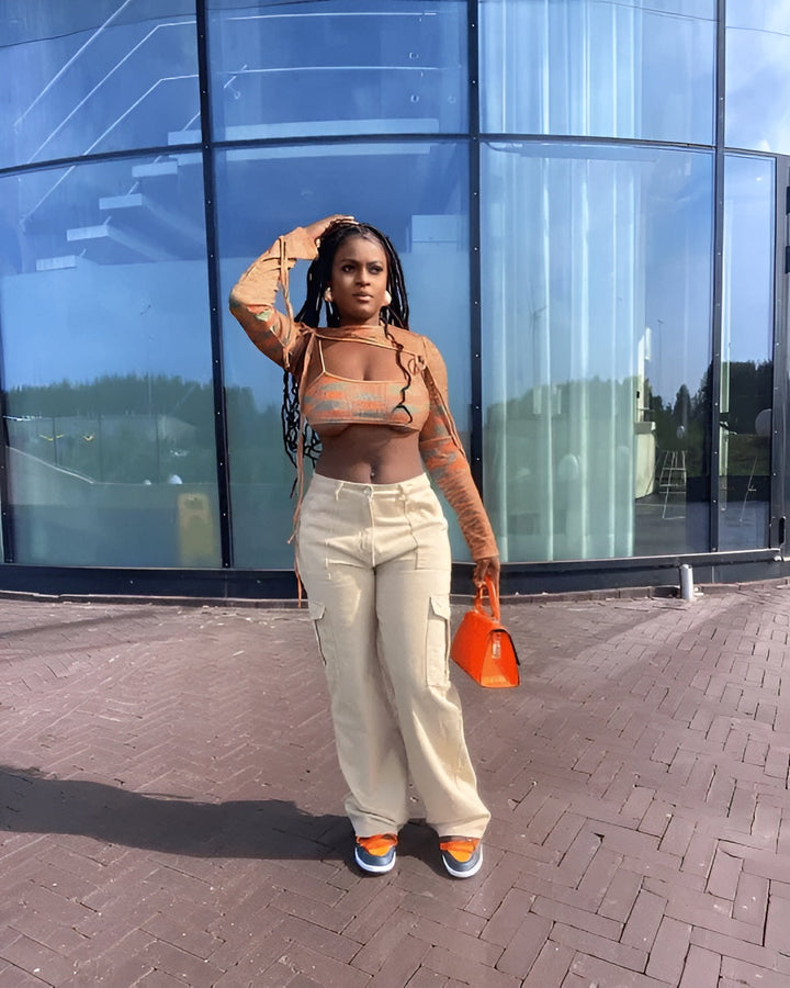 Frau in orangefarbenem Crop-Top, beigen Cargo-Hosen, mit orangefarbener Tasche vor Glasgebäude.