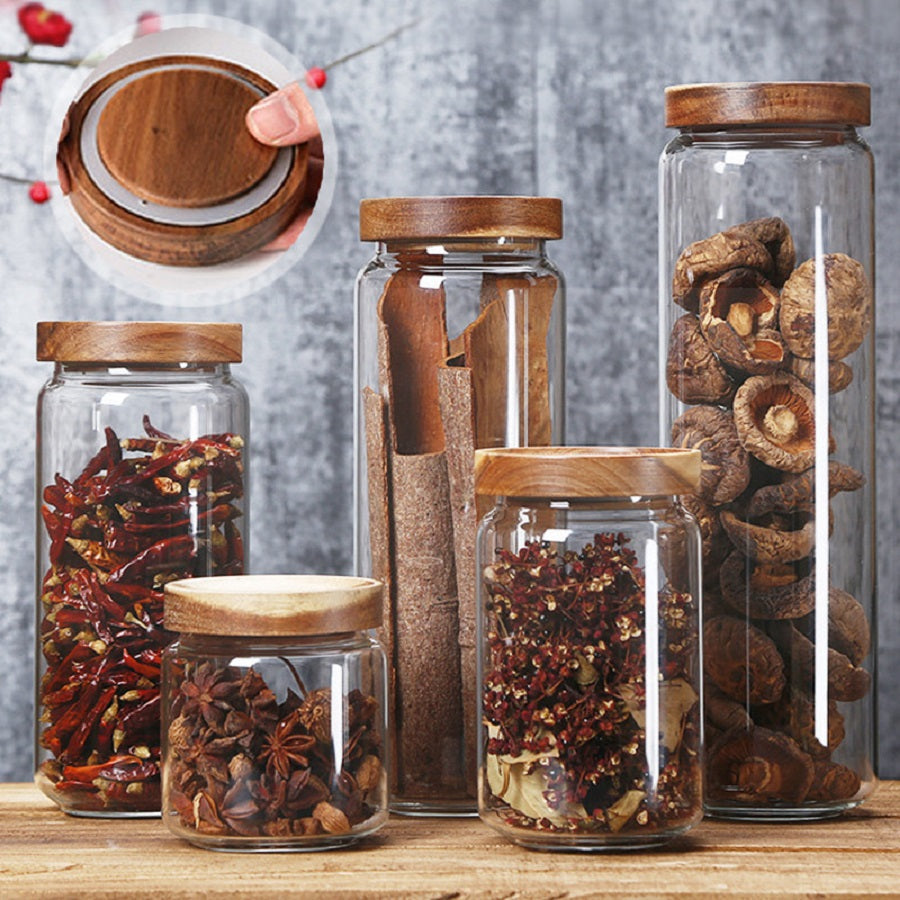 Fünf Glasbehälter mit Holzdeckeln, gefüllt mit Gewürzen und getrockneten Pilzen, auf Holztisch.