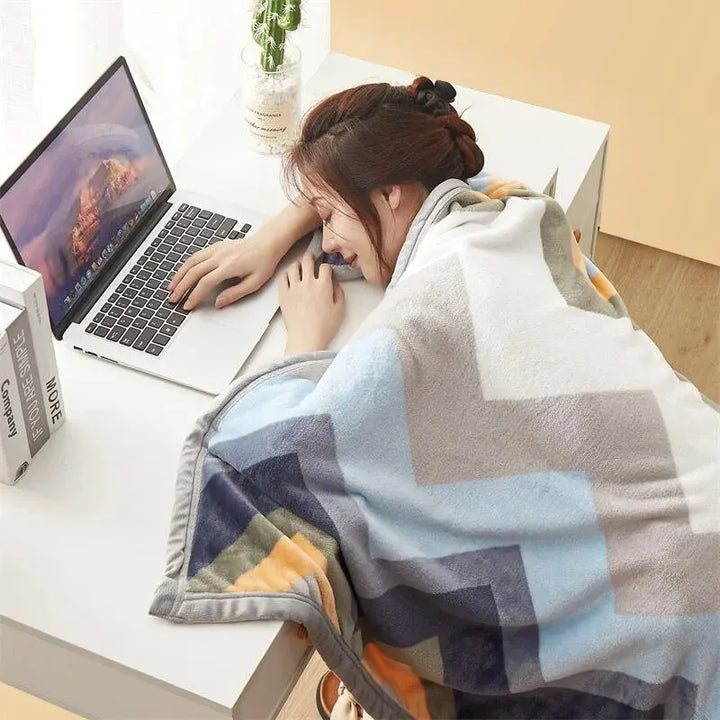 Frau schläft auf Schreibtisch mit Laptop, eingewickelt in geometrische Decke, Homeoffice-Szene.