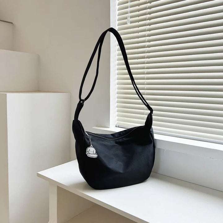 Schwarze Umhängetasche aus Nylon, modern, minimalistisch, auf weißem Tisch vor Jalousie.