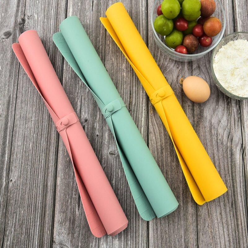Drei bunte Silikon-Backmatten auf Holztisch, rosa, grün, gelb, neben Eiern und Obst.