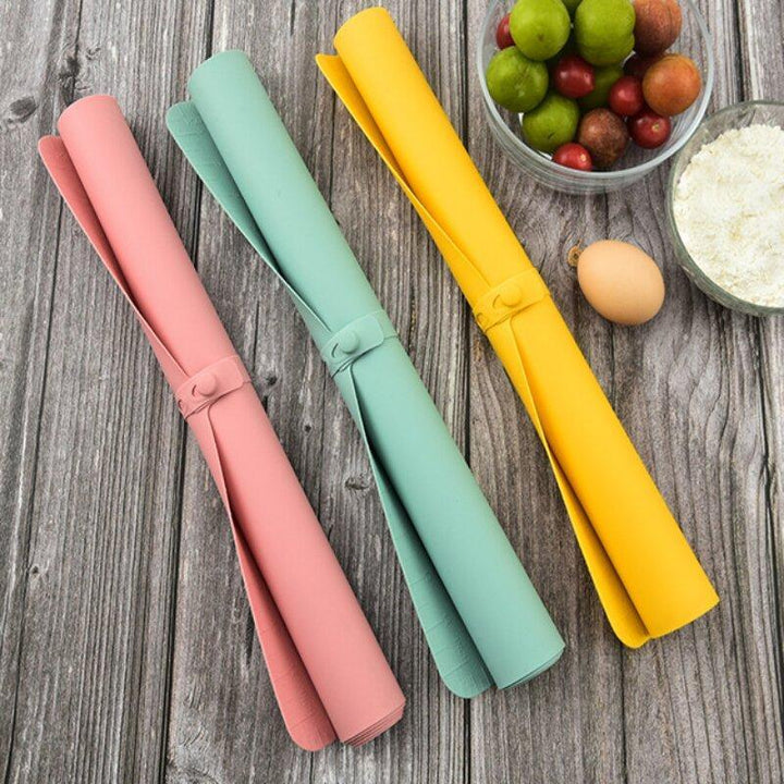 Drei bunte Silikon-Backmatten auf Holztisch, rosa, grün, gelb, neben Eiern und Obst.