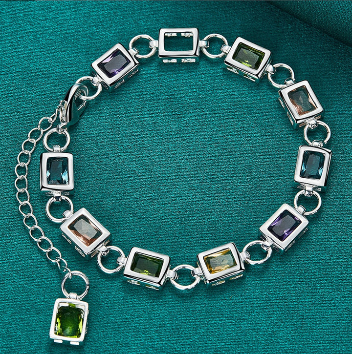 Silberarmband mit rechteckigen Edelsteinen, grün und lila, auf grünem Hintergrund.