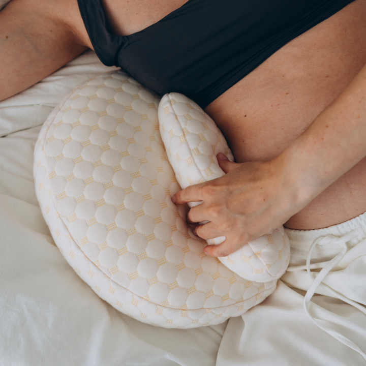 Frau mit weißem Wärmekissen auf Bauch, Linderung Menstruationsschmerzen, gemütliches Schlafzimmer.