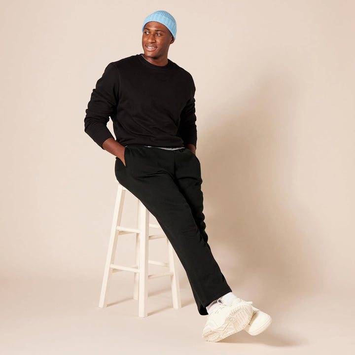 Mann in schwarzem Outfit auf weißem Hocker, blaue Mütze, weiße Sneaker, beige Hintergrund.