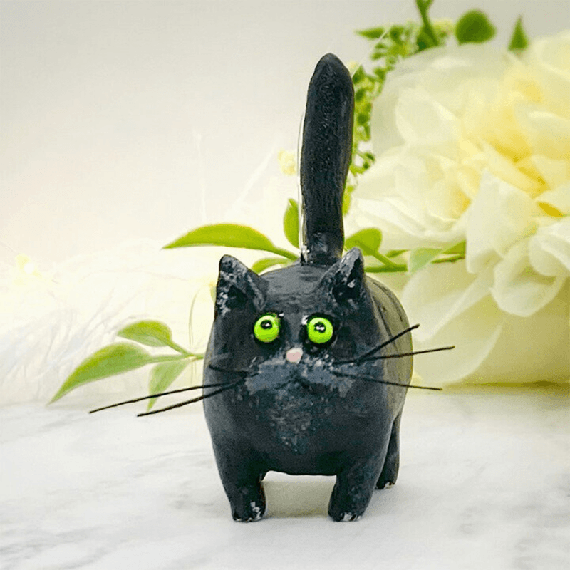 Schwarze Katzenfigur aus Keramik mit grünen Augen, handgefertigt, Dekoration, vor weißen Blumen.
