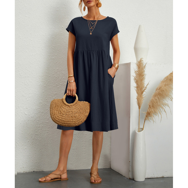 Frau in dunkelblauem Baumwoll-Leinen-Kleid, Boho-Stil, mit Strohtasche und Sandalen.