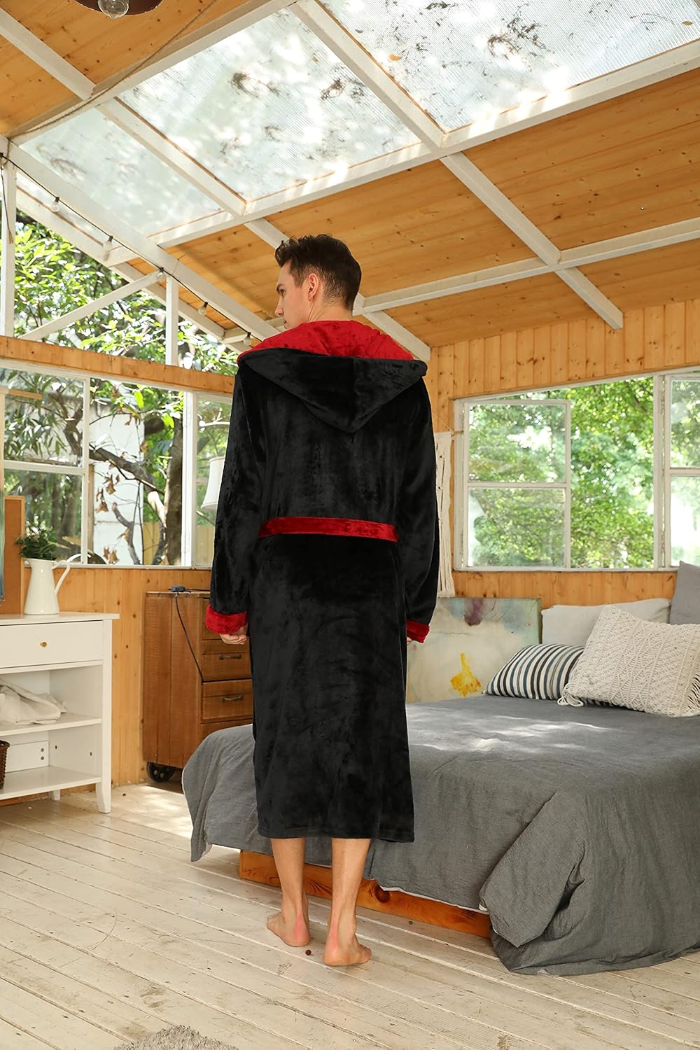 Mann im schwarzen Bademantel mit rotem Futter, Kapuze, in hellem Schlafzimmer.