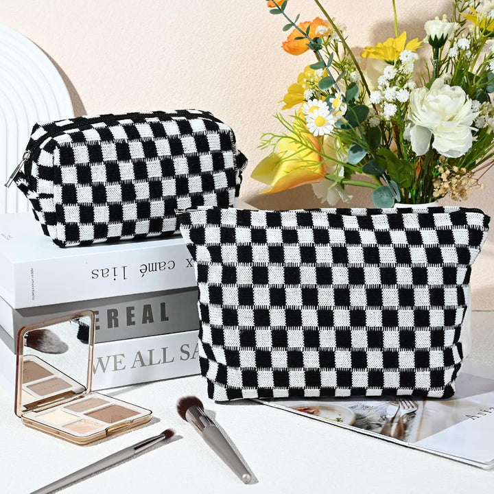 Schwarz-weiß karierte Kosmetiktaschen auf Büchern, mit Blumen und Make-up-Pinsel im Hintergrund.