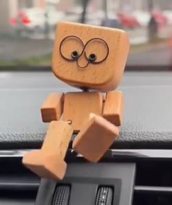 Detail dřevěné figurky s velkýma očima, která sedí na palubní desce auta a nohy jí visí dolů. 6