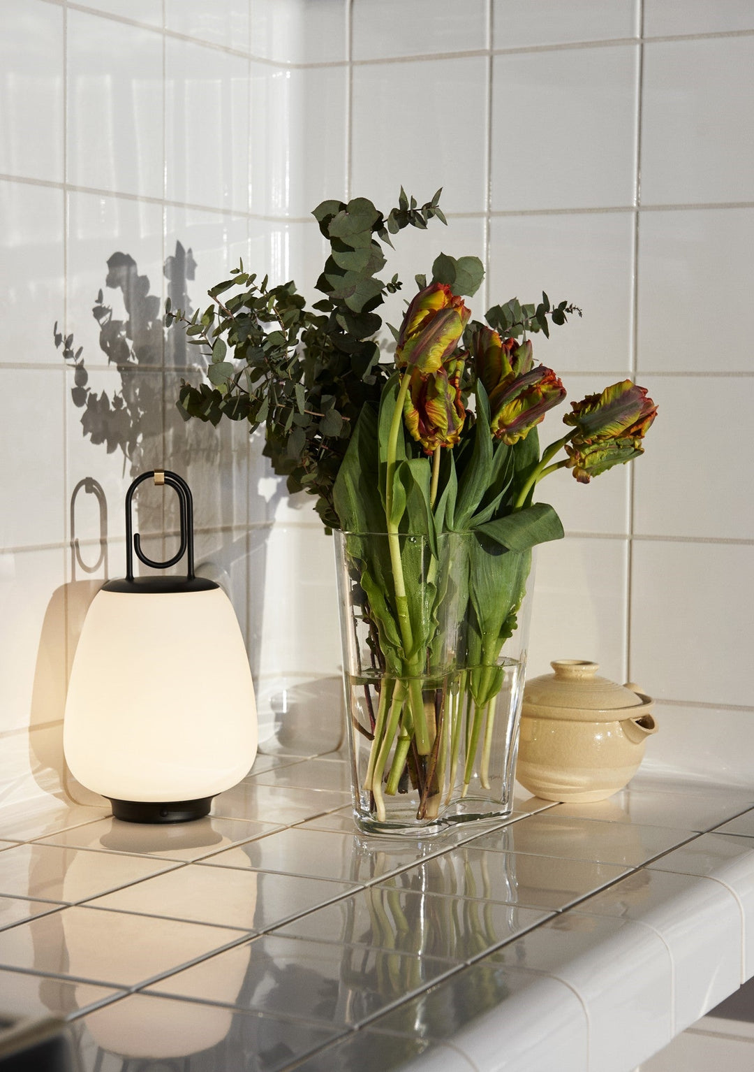 Vase mit Tulpen und Eukalyptus auf weißem Fliesentisch neben moderner Tischlampe.
