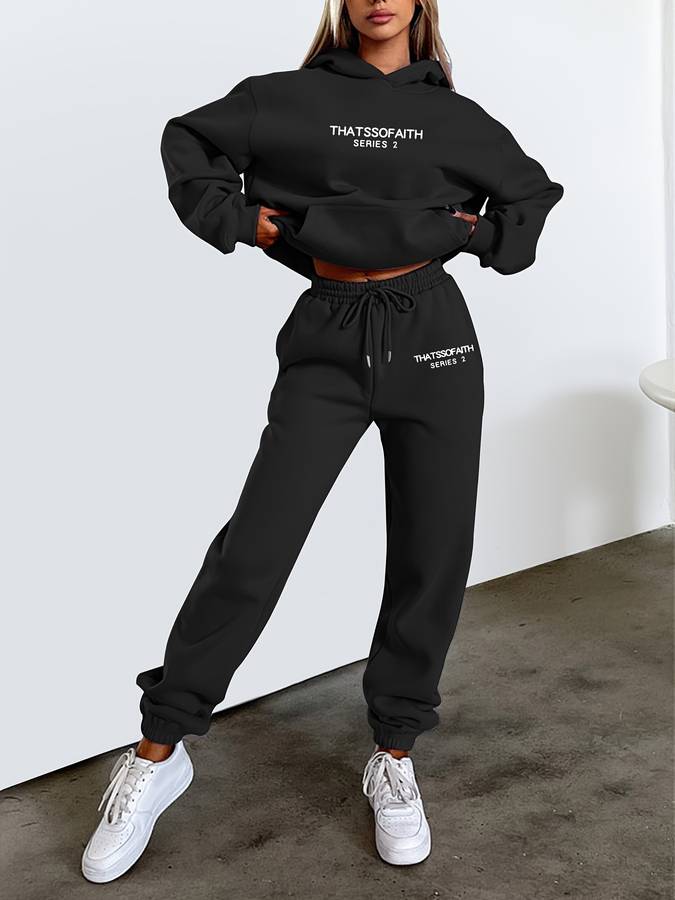 Frau in schwarzem Hoodie und Jogginghose, THATSSOFAITH-Logo, weiße Sneaker, lässige Mode.